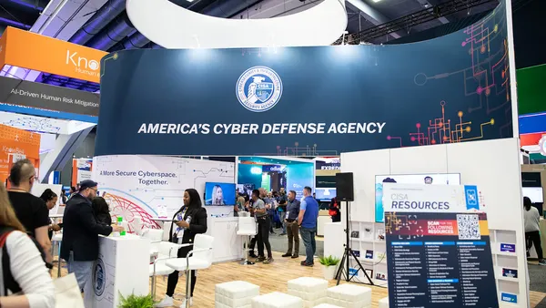A banner bearing the eagle-and-shield logo of the Cybersecurity and Infrastructure Security Agency and reading "America's Cyber Defense Agency" hangs over a conference booth that contains chairs, posters, speakers, and plants.