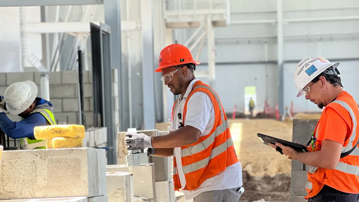 Construction workers in safety gear collaborating on a building site, with one using a tablet and others laying bricks.