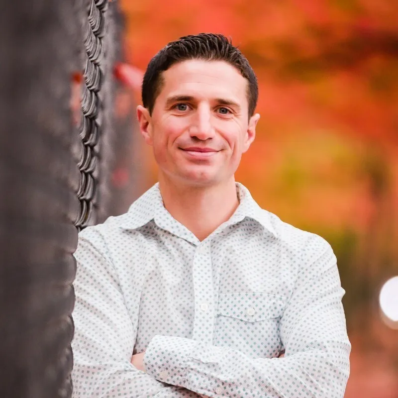 A headshot of a man leaning on a chain link fence against a background of autumn leaves