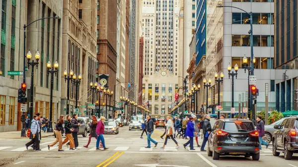 People walk across a city street at a crosswalk. Tall buildings and streetlights line the road,