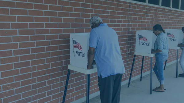 3 People voting at individual voting booths at a voting station outside of a brick building.