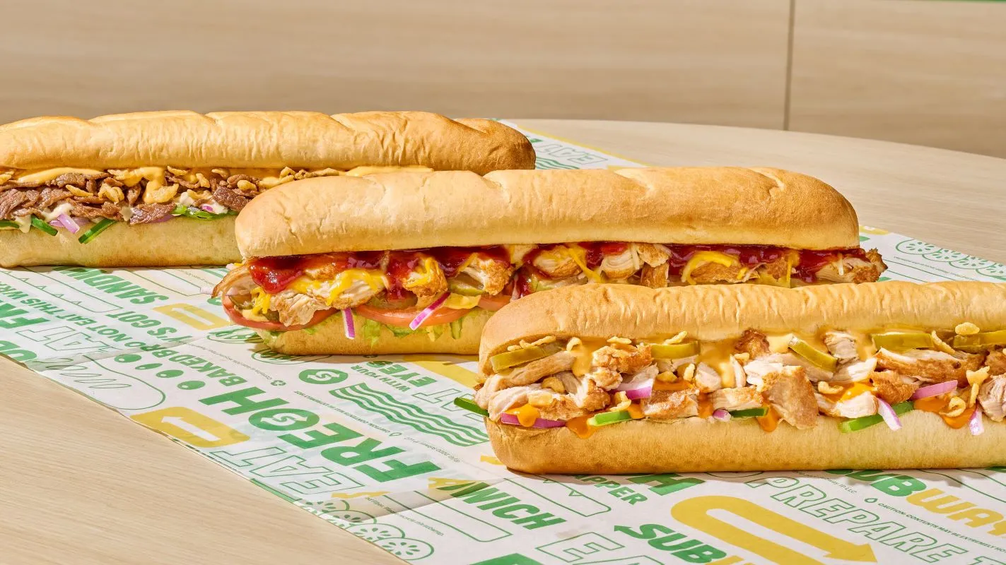 Three Subway sandwiches on a table on top of Subway wrapping paper.