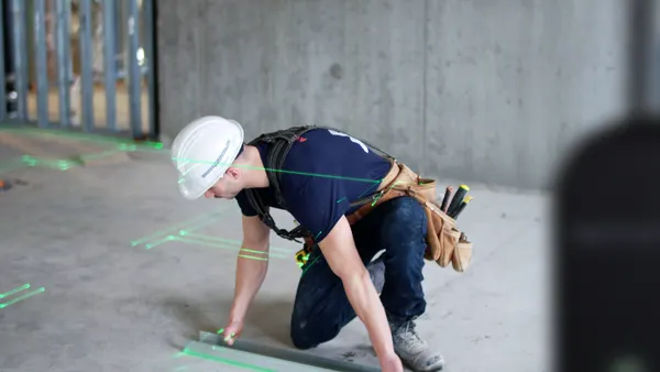 A person with a white hardhat kneels on a floor while a machine projects green light onto the ground.