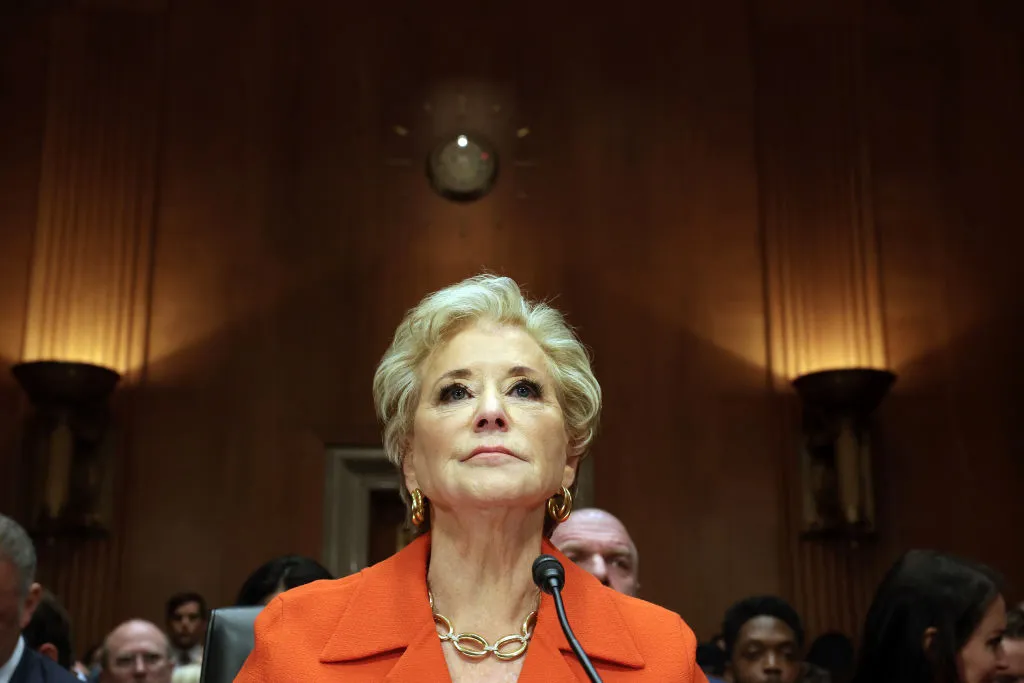 A person is photographed from the front and below. Only the person&#x27;s head, neck and shoulders can be seen. They are in a room with wood paneling. Others are seated behind the person.