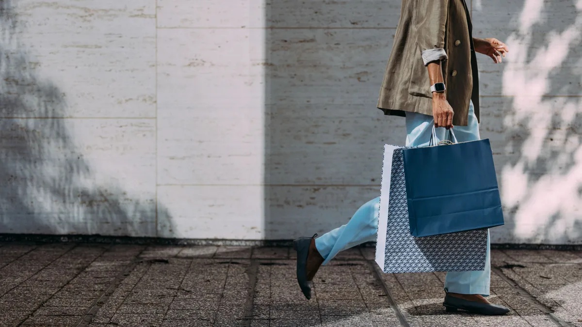 A person walks down a city street wearing jeans and carrying two shopping bags.