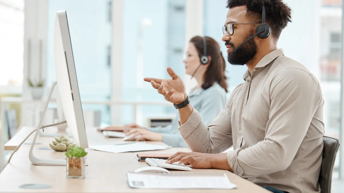 Picture of a man and a woman working at two computer stations in an office with headsets on.