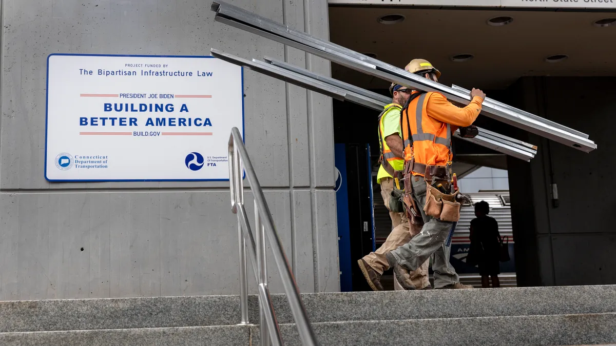 Two construction workers carry materials past a sign promoting infrastructure.