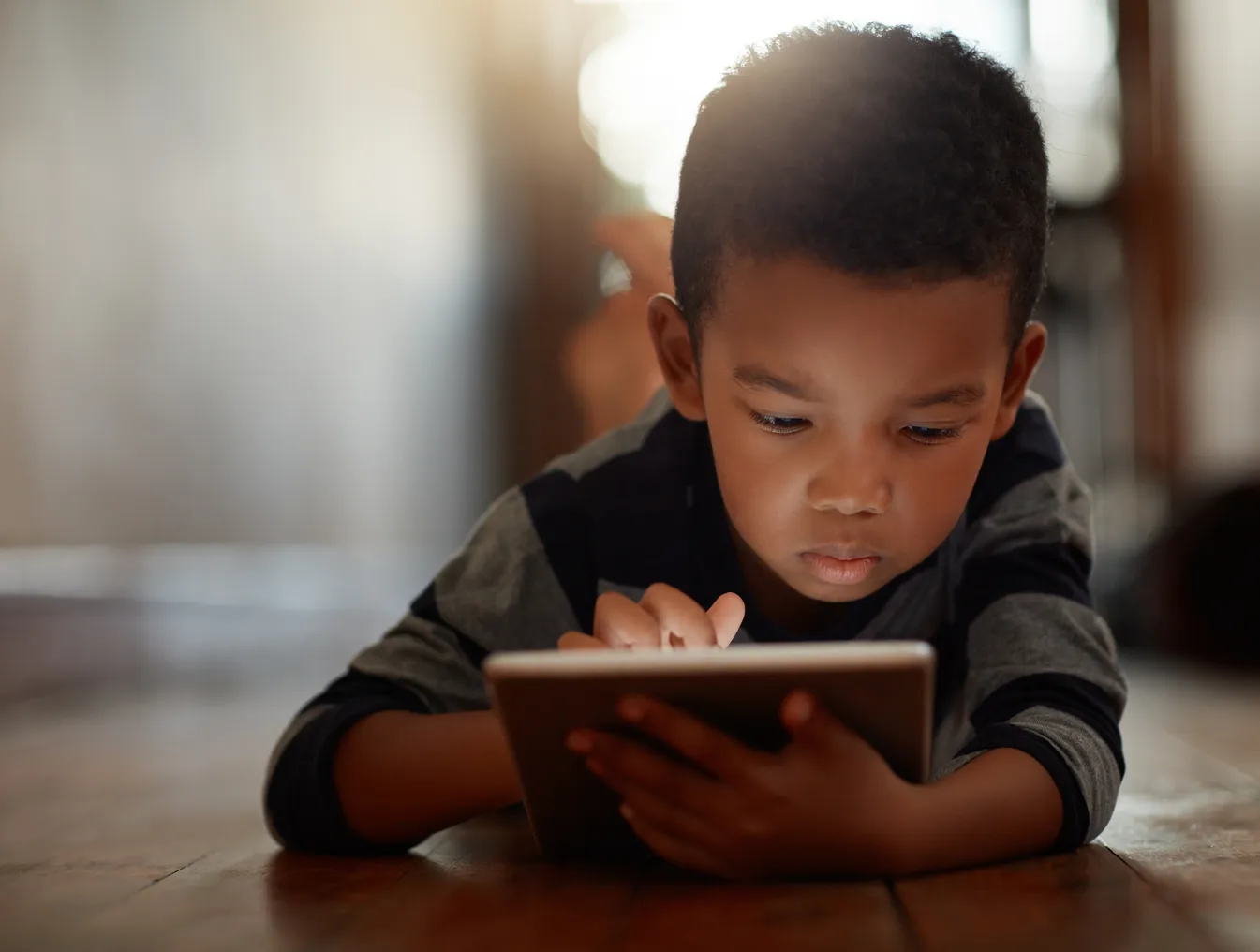 A young student lies with stomach on wood floor with a tablet . One hand is on the screen of the tablet.