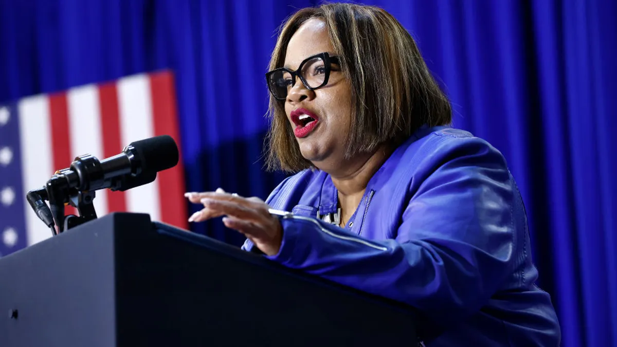 A Black woman in a blue jacket and statement glasses speaks in front of a blue background with an American flag