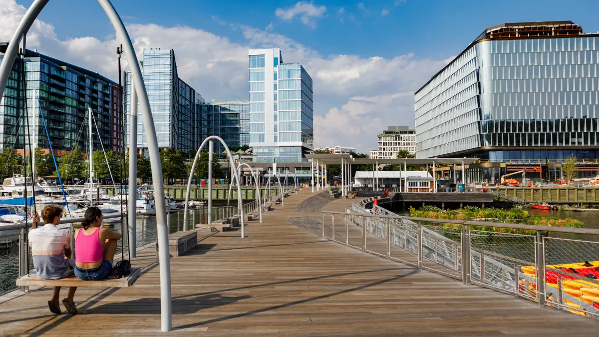 Two people sit on a swing on the left of the photo with a wooden boardwalk down the middle and large blue and white buildings in the background on a sunny day.