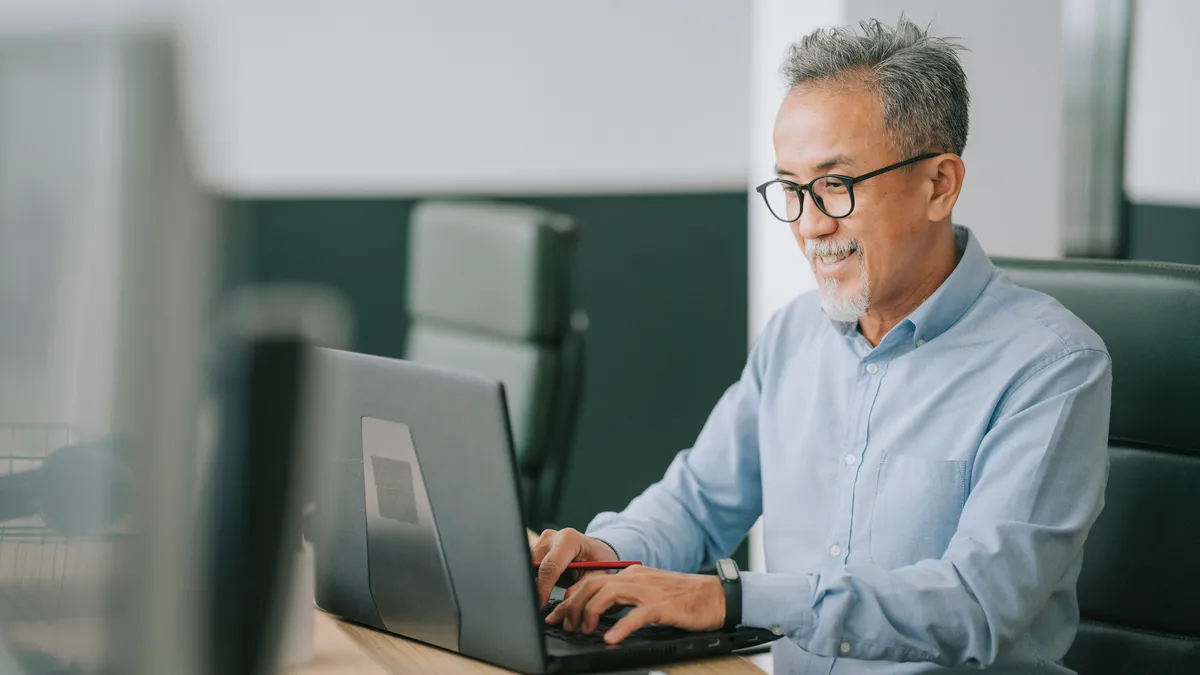 Asian senior man with facial hair using laptop typing working in office open plan