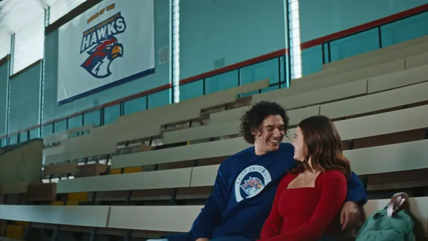A boy and a girl sit in hockey bleachers. The boy, who is in a blue sweatshirt, has his arm around the girl, who is in red.