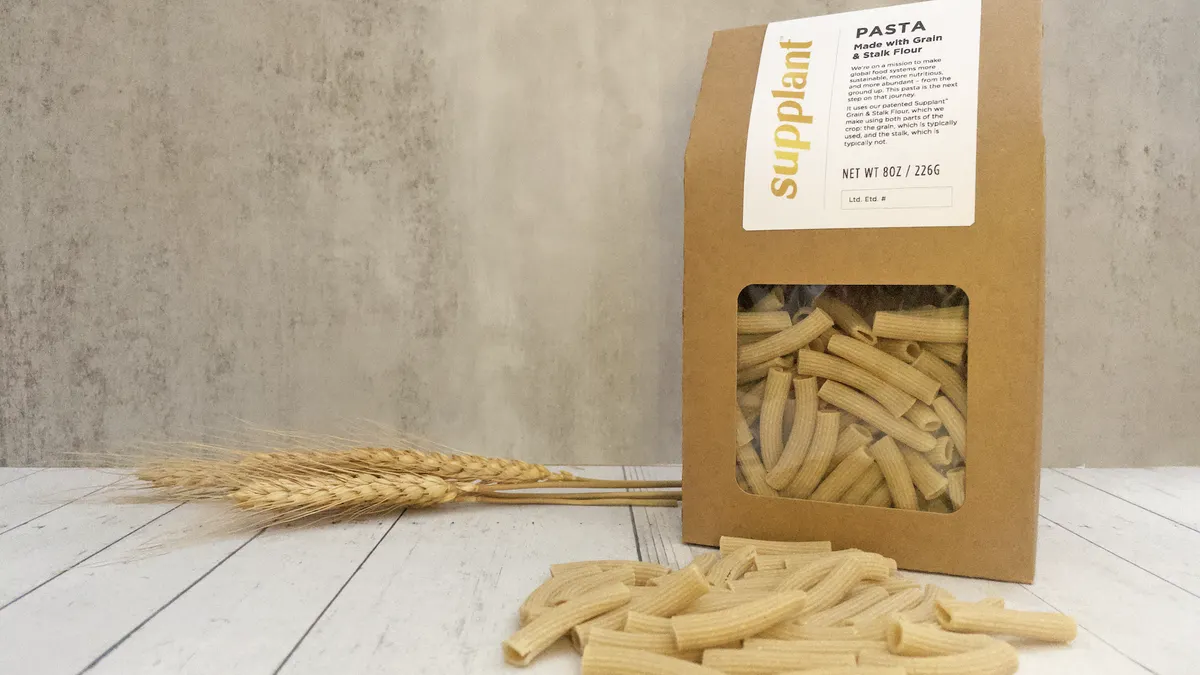 A brown paper package of Supplant Grain & Stalk Pasta sits to the right on a white wooden table with noodles in front of it and wheat stalks behind. The background is marbled gray.