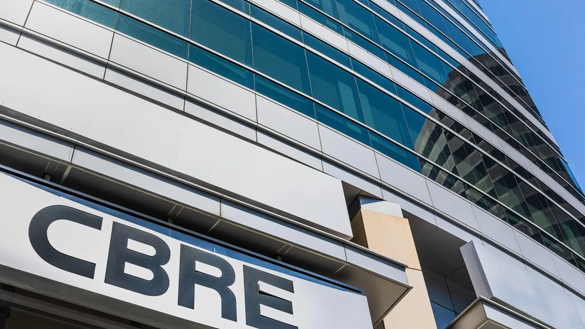 Upward view of a modern glass and steel office building with the CBRE logo prominently displayed on a sign at the entrance. The structure features reflective blue-tinted windows, and another curved building is reflected in the glass façade.