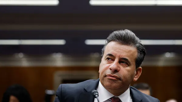 A person in a dark business suit sits at a desk with a microphone.