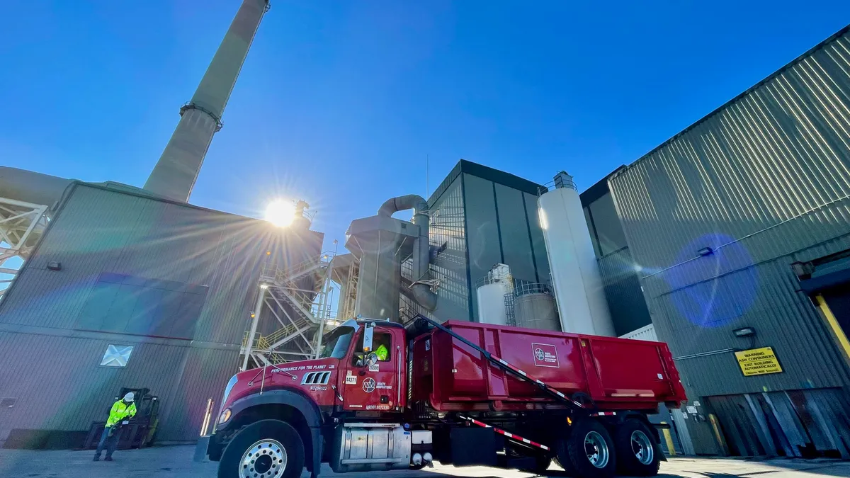 Red waste truck in front of industrial facility with smokestack in the background