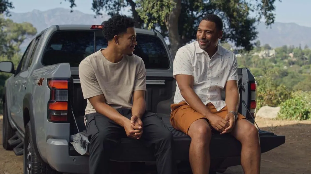 Two men smile and talk in the bed of a grey truck parked in an outdoor space.