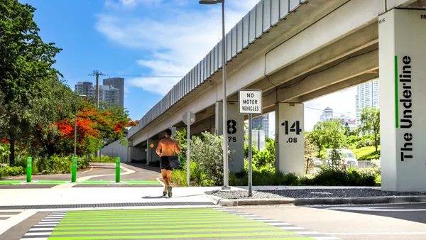 A person in shorts and sneakers runs along a tree-lined path next to an elevated track.