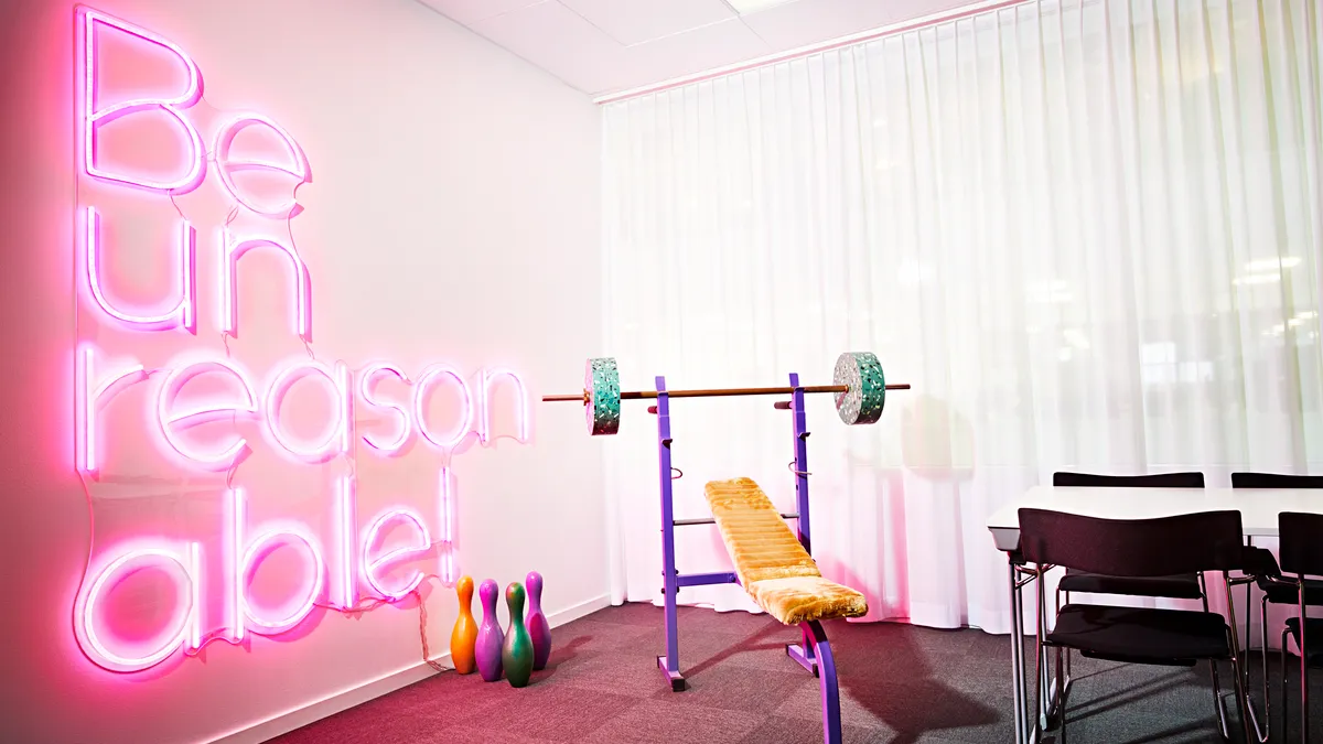 Room with a neon sign that says 'Be Unreasonable' with weights and a desk.