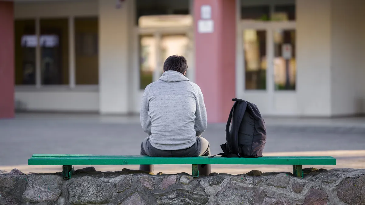 A student is sitting outside a school building on a bench with their back toward the camera. A backpack sits next to the person on the bench.