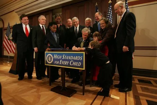 A person is sitting at a desk on a stage. People, including children are gathered around and behind the person at the desk who is writing something. A sign in from the desk says &quot;High Standards For Every Child&quot;