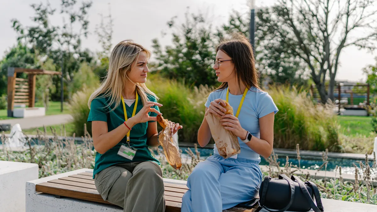 Two young female nurses in scrubs taking a lunch break in the park.