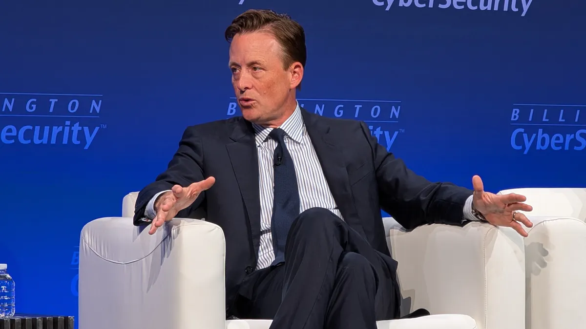 A man wearing a dark blue suit and tie gestures while sitting in a plush white chair in front of a backdrop that reads "Billington Cybersecurity"