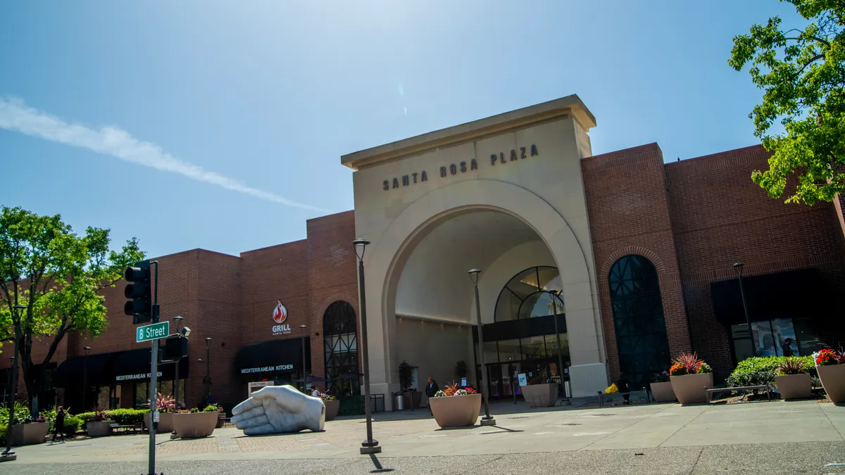 The arched entrance to a building reads "Santa Rosa Plaza," with trees, potted flowers and a sculpture of a hand in the foreground, and cloud-streaked blue sky in the background.