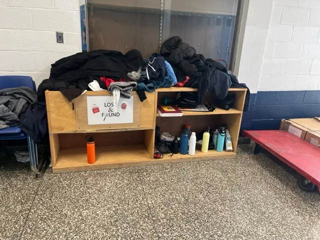 A short wooden bin with shelves displays coats, jackets, water bottles, sneakers, notebooks and more. The bin is inside a building against a brick wall.