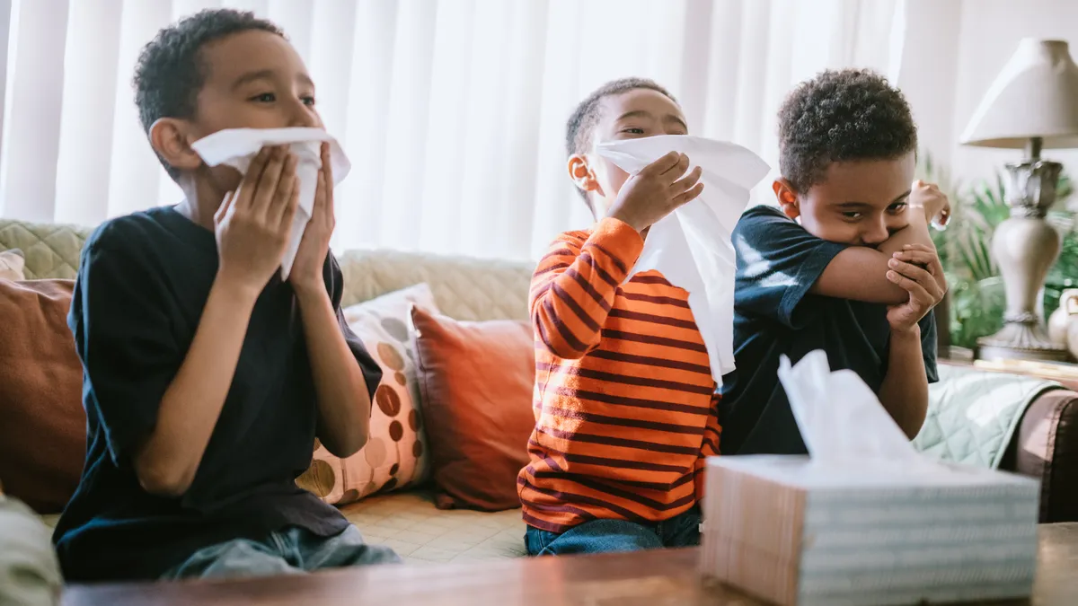 Three boys cough and sneeze and wipe their faces with tissues standing between a sofa and coffee table in a living room.