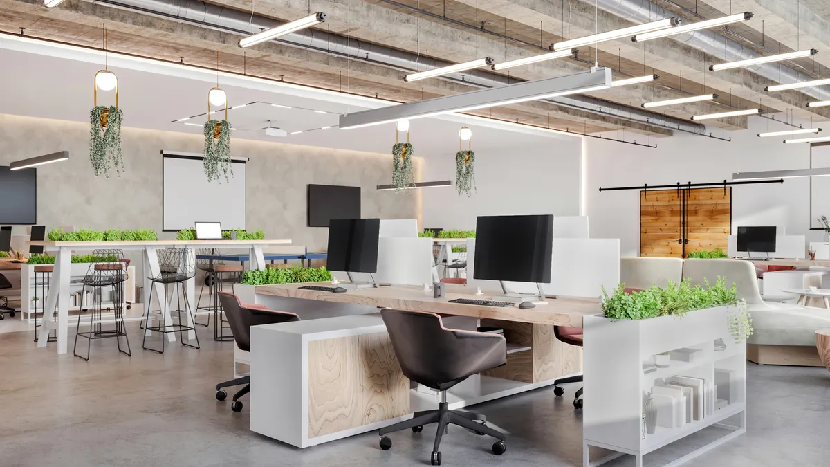 A modern open-plan office space interior featuring desks, computers, armchairs, a shelf, a plant a concrete ceiling and LED lights.