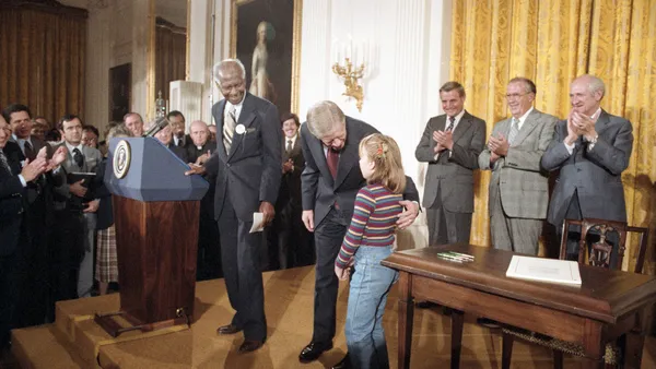 Adults stand around and on a stage that has a podium and desk. An adult is leaning down toward a child with their arm around the child.