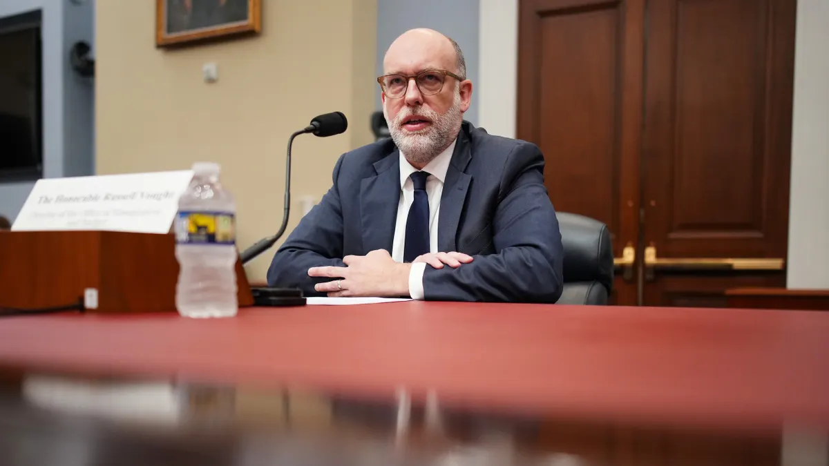 A bald man in a dark suit and tie sits at a desk in front of a microphone.