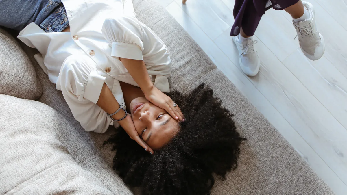 An overhead shot of a Black woman clutching her head in distress on a couch