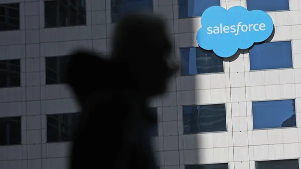 A corporate building bearing the logo of Salesforce in the background as the shadow of a person wearing a hoodie remains out of focus in the foreground