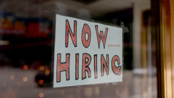 A handwritten "now hiring" sign hangs in a shop window.