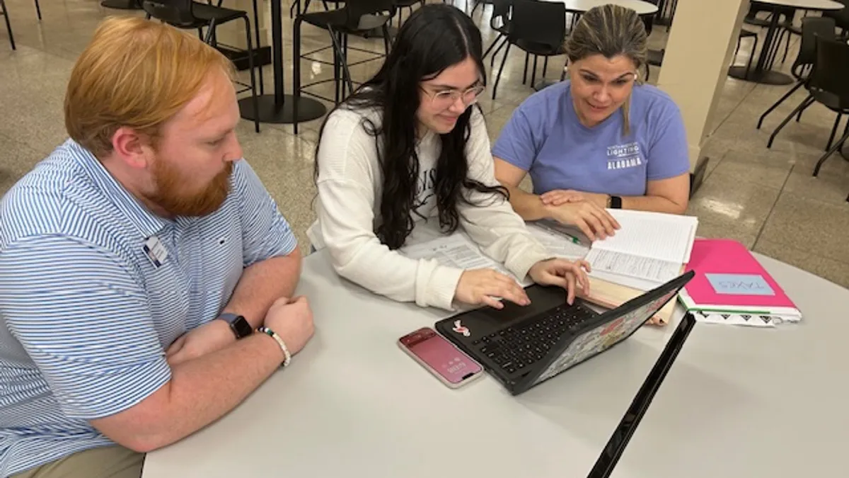 Three people sit at a table. The person in the center is using a laptop.