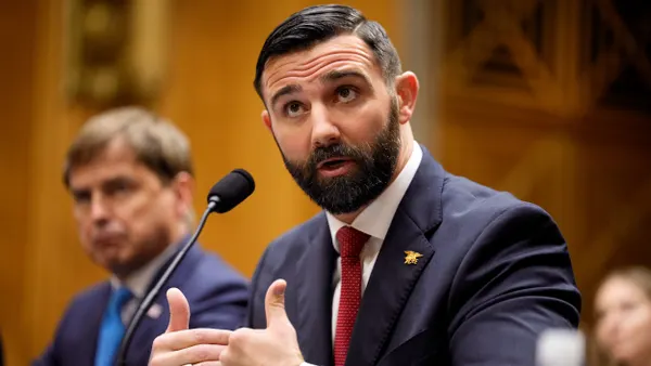 A person with a beard wearing a blue suit talks into a microphone.