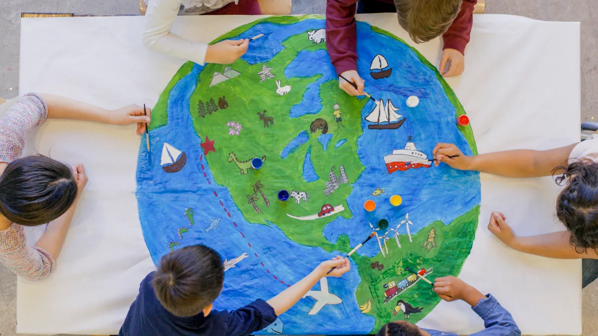 Photo looks down on several young students seated at a table. Students are drawing on a picture of continents and oceans with drawing of ships and planes.