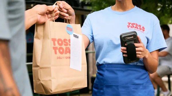 Food worker in a blue shirt hands a takeout bag to a customer.