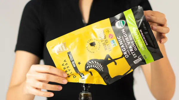 A person uses a pouch containing olive to refill a bottle with yellow and black Terra Delyssa olive oil branding on the outside.