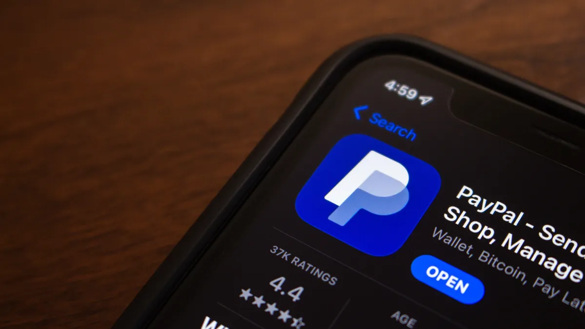 The PayPal app is displayed in the Apple App Store on an iPhone screen, showcasing the app's icon and download option; the phone is on a brown wooden surface. 