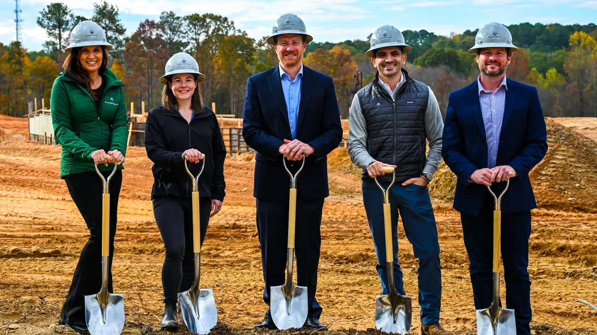 Five people stand together on a dirt surface with shovels and hard hats.