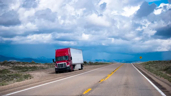 A refrigerated tractor-trailer is stuck on the side of a deserted road with a mountain range in the background.