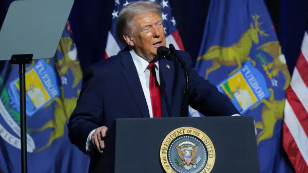 President Donald Trump, wearing blue suit and red tie, speaks into microphone at podium with presidential seal in front of several U.S. flags.