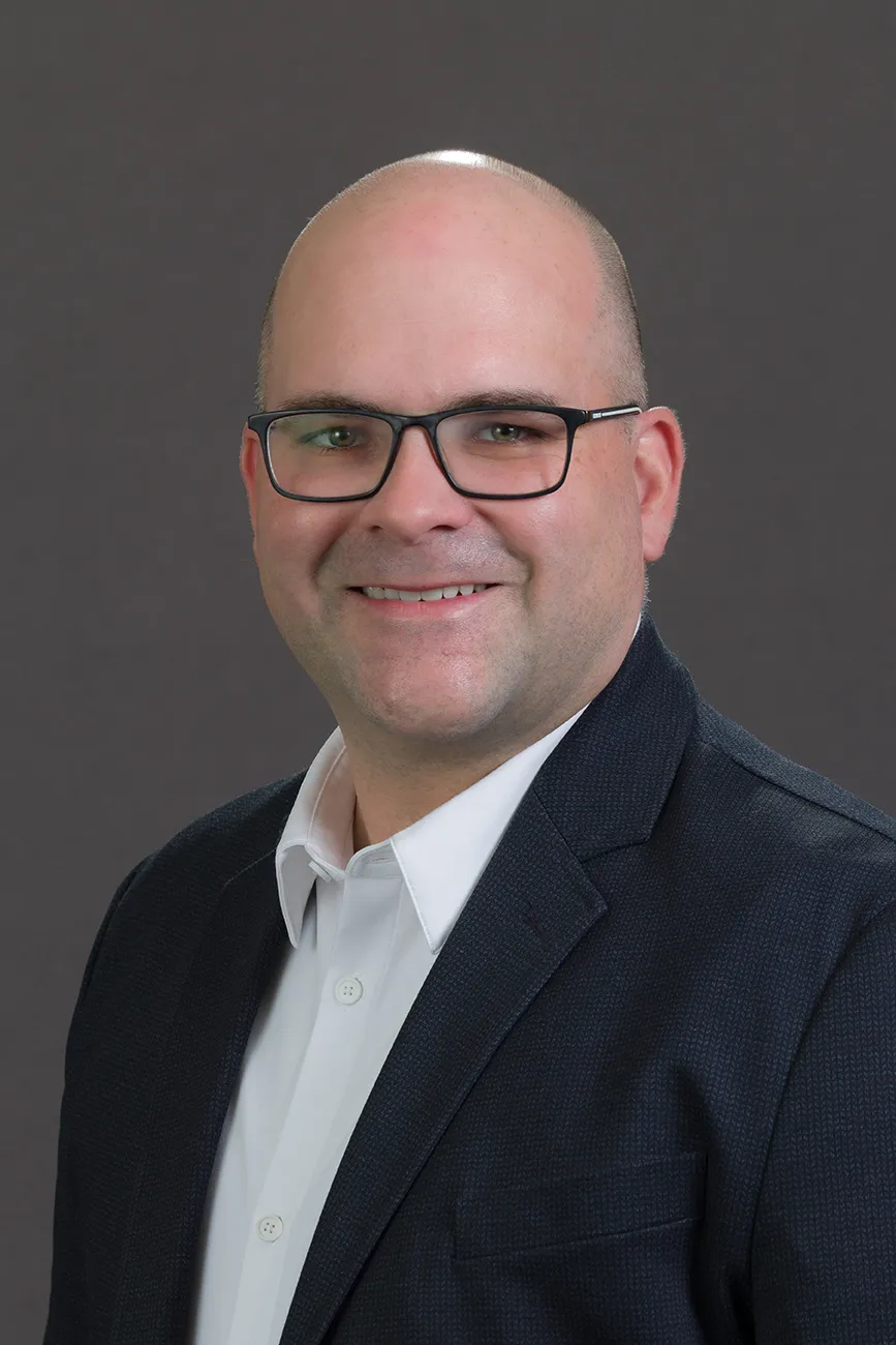 A man wearing a suit and glasses in front of a neutral background.