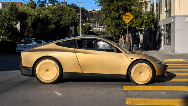 A sleek 2-door bronze color vehicle crosses an intersection with trees and buildings in the background.