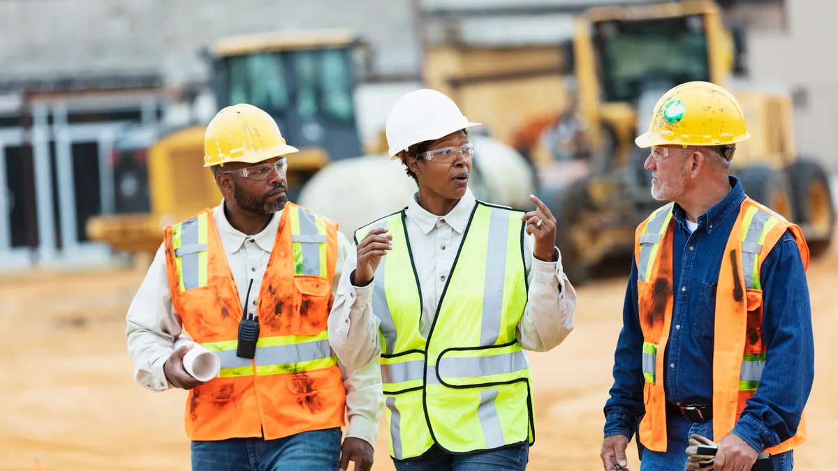 A multiracial group of three construction workers, led by an African-American woman, at a construction site. The woman, in her 40s, is walking between two men in their 50s, talking and looking toward the Hispanic man.