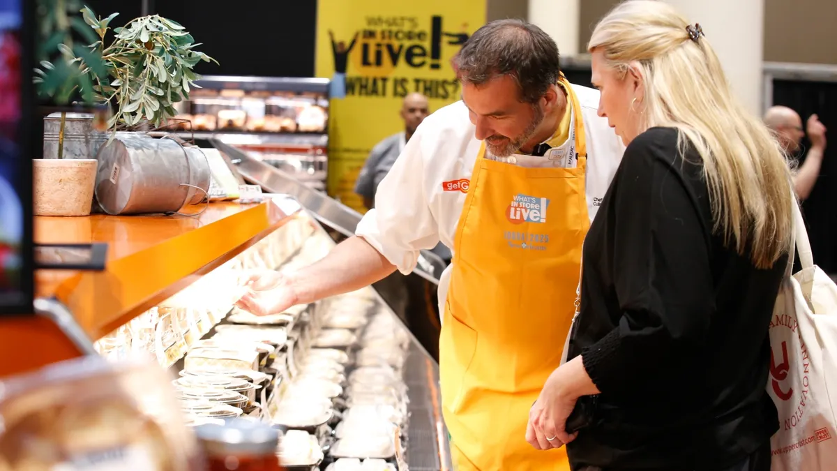 Person in an orange apron shows food products to someone at a grocery trade show.