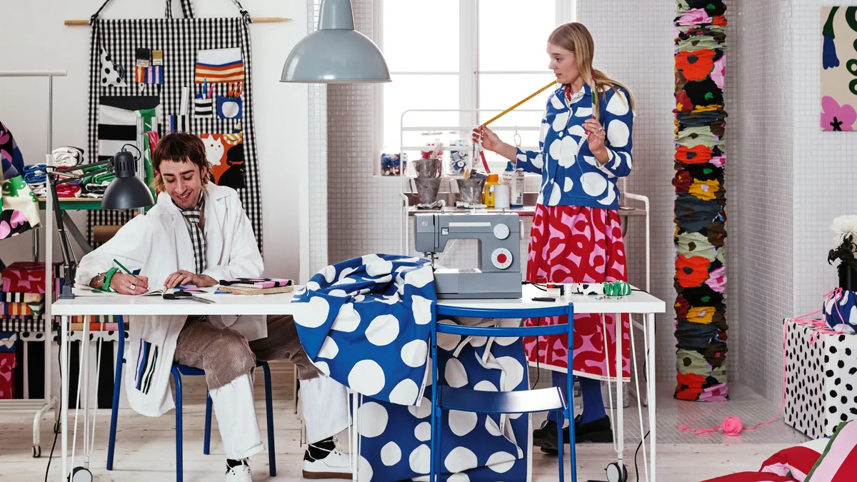 Two people are in a white room surrounded by color prints and fabrics.